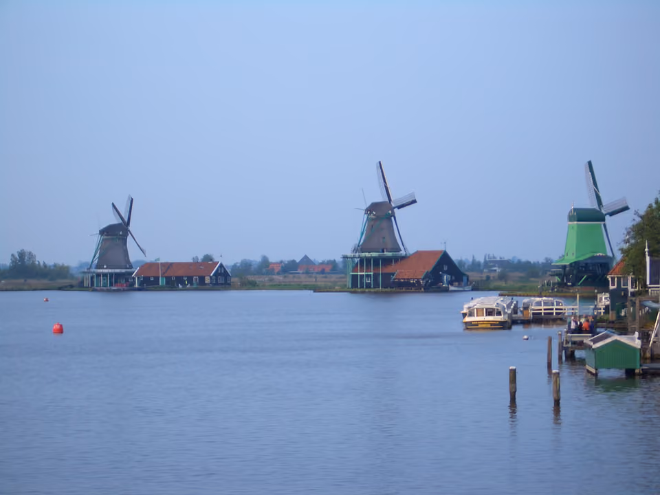 Moinhos de vento históricos de Zaanse Schans à beira de um canal, com céu nublado e um pequeno barco atracado na margem