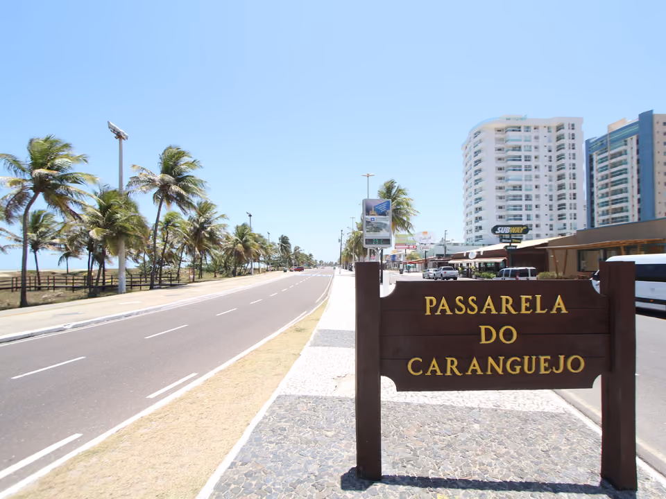 Placa da Passarela do Caranguejo na orla de Aracaju, com coqueiral e prédios ao fundo