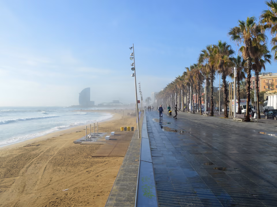 Praia e calçadão da Barceloneta em Barcelona, com as palmeiras à direita, a faixa de areia à esquerda e o edifício em forma de vela do Hotel W na ponta, na bruma da manhã