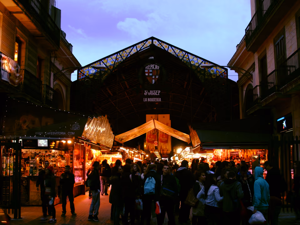 Entrada do Mercado da Boqueria na La Rambla de Barcelona ao entardecer, com o letreiro de vitral colorido e o brasão, a estrutura de ferro e as bancas iluminadas ao fundo