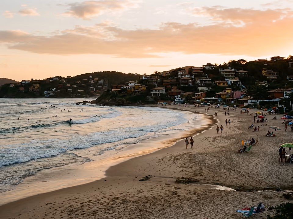Arco extenso de areia de Geribá com silhuetas de surfistas ao longe e casas nas colinas
