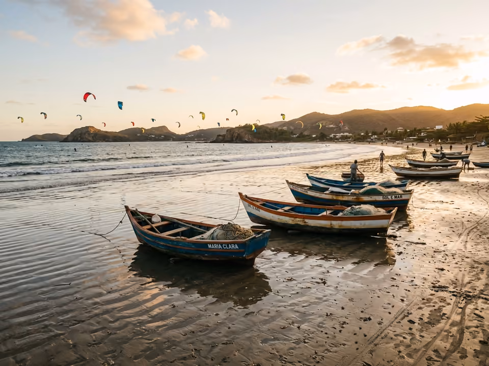 Maré baixa em Manguinhos com kites ao longe e barcos de pesca na areia molhada refletindo o céu