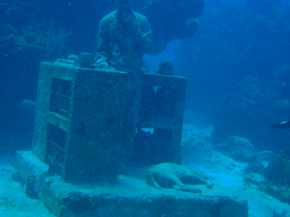 Escultura submersa de uma figura humana diante de uma cômoda, coberta de coral, no Museu Subaquático de Arte de Cancún