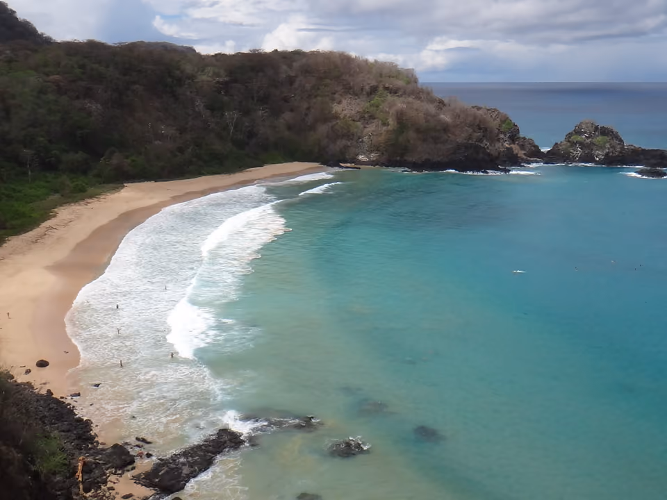 Baía do Sancho em Fernando de Noronha vista do mirante, a meia-lua de areia clara ao pé de uma falésia coberta de mata, com o mar em faixas de verde e azul
