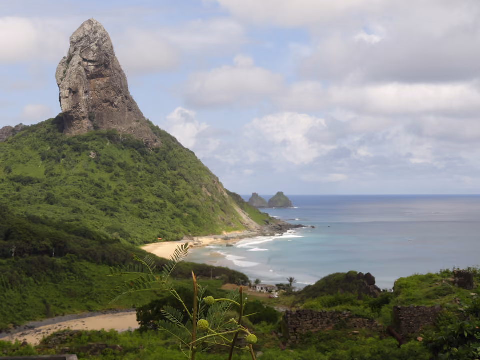 O Morro do Pico, a torre de rocha de 321 metros de Fernando de Noronha, erguendo-se sobre a encosta verde e a Praia da Conceição, com o Morro Dois Irmãos no horizonte