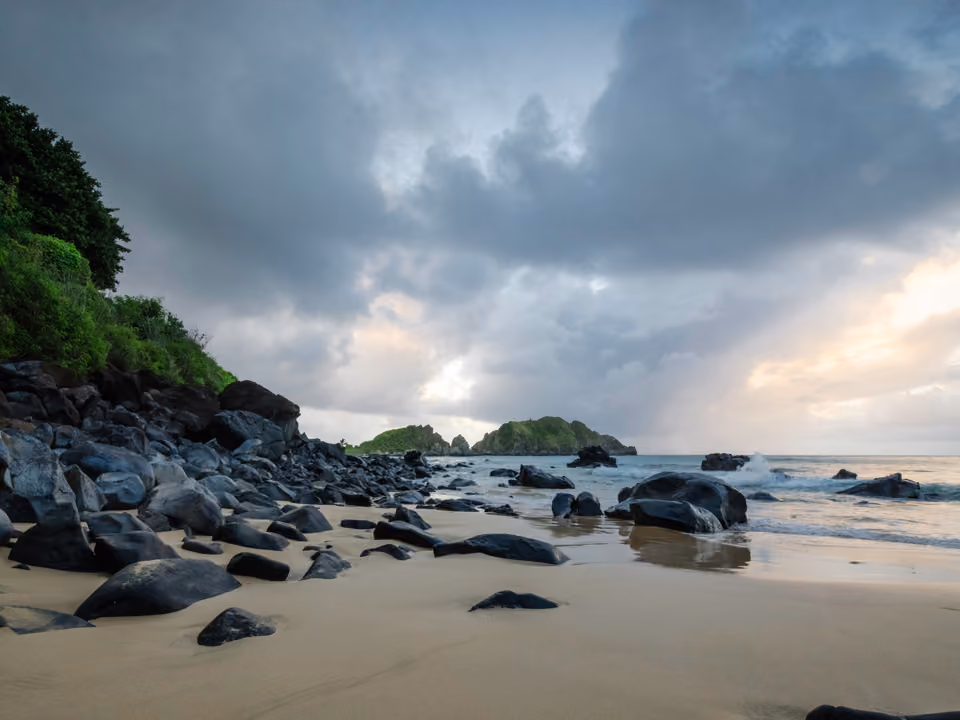 Praia do Cachorro em Fernando de Noronha, pequena faixa de areia com grandes blocos de rocha vulcânica escura, ao pé de uma falésia verde, sob céu de fim de tarde nublado