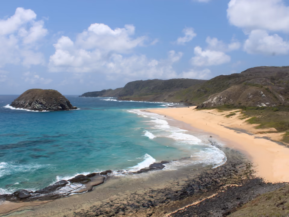 Praia do Leão em Fernando de Noronha, longa faixa de areia quase deserta em curva, com a ilha rochosa do Leão em frente e o mar aberto com arrebentação