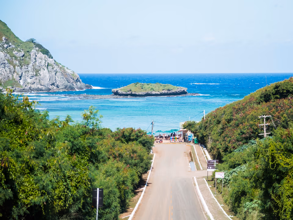 A enseada calma e rasa da Praia do Sueste em Fernando de Noronha, com um pequeno ilhote e o posto de controle no fim da estrada que desce à praia