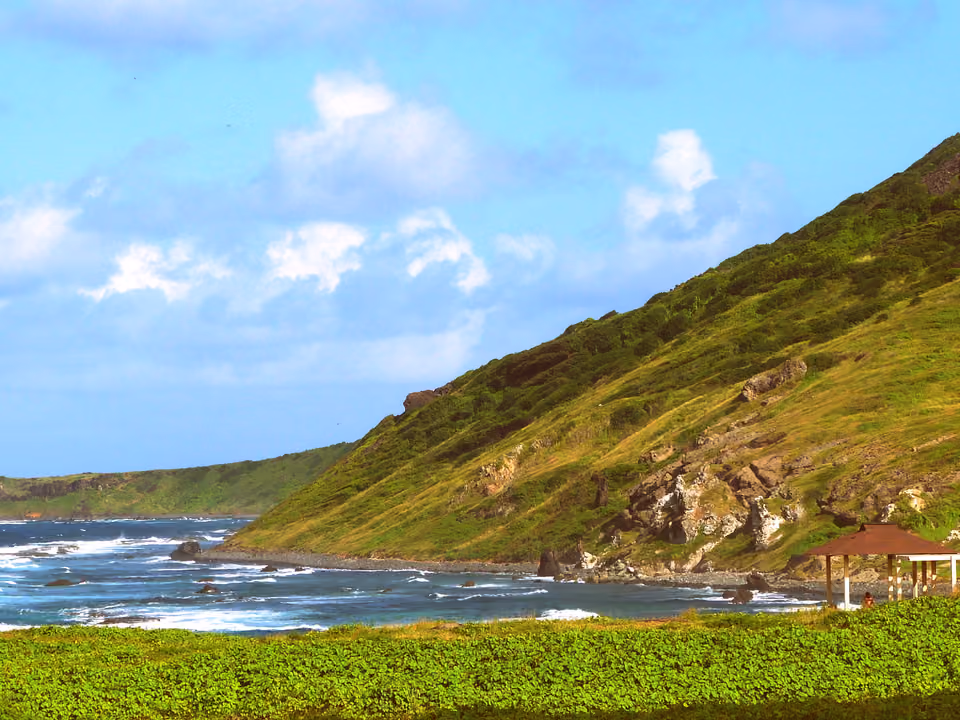A costa rochosa da Praia do Atalaia em Fernando de Noronha na maré alta, encosta verde descendo ao mar e um abrigo de palha do controle da trilha à direita
