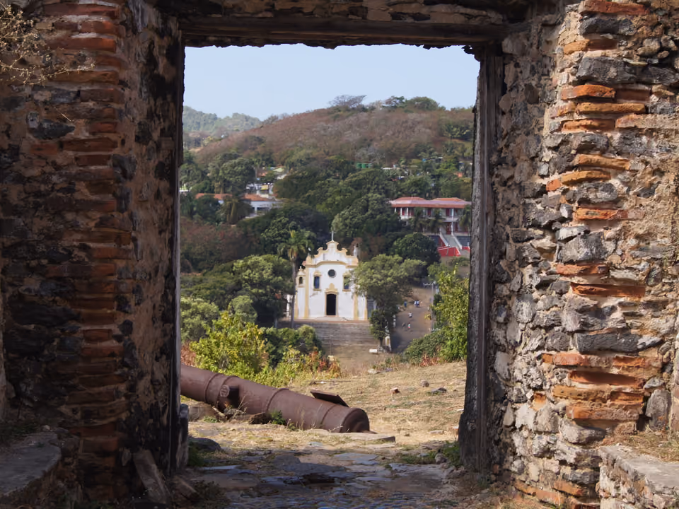 A Igreja de Nossa Senhora dos Remédios, capela colonial branca de Fernando de Noronha, emoldurada por uma porta em ruínas do forte, com um canhão antigo no primeiro plano e a Vila ao fundo