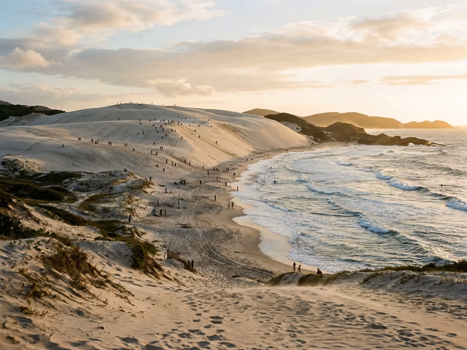 Dunas brancas da Joaquina com silhuetas distantes de sandboard e ondas quebrando