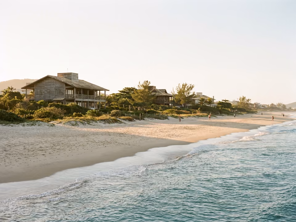 Faixa larga e plana de areia de Jurerê com mar calmo turquesa e casas elegantes atrás da vegetação