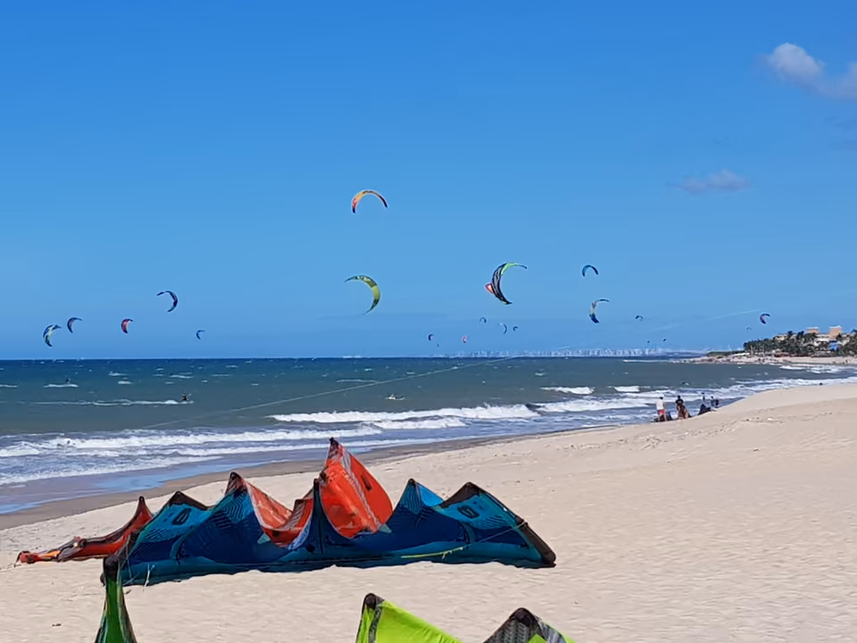 Dezenas de pipas de kitesurf coloridas no céu sobre o mar de Cumbuco, com equipamento de kite na areia em primeiro plano