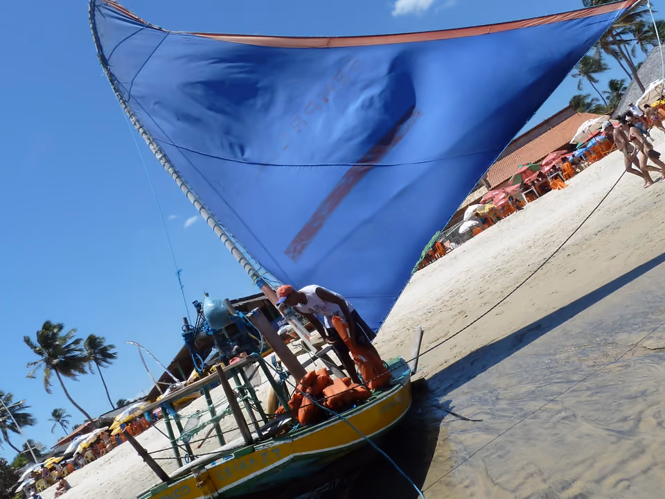 Jangada tradicional cearense com vela azul, colete salva-vidas a bordo e barracas de praia ao fundo