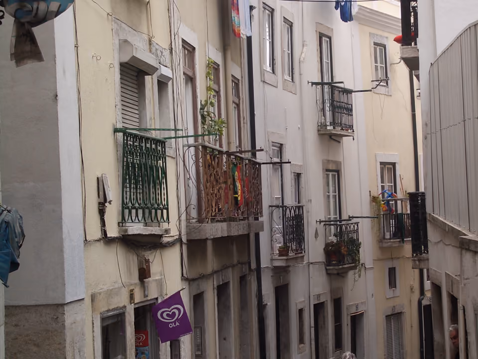 Rua estreita de Alfama em Lisboa, com casario claro, varandas de ferro, roupa no varal e uma bandeira de Portugal