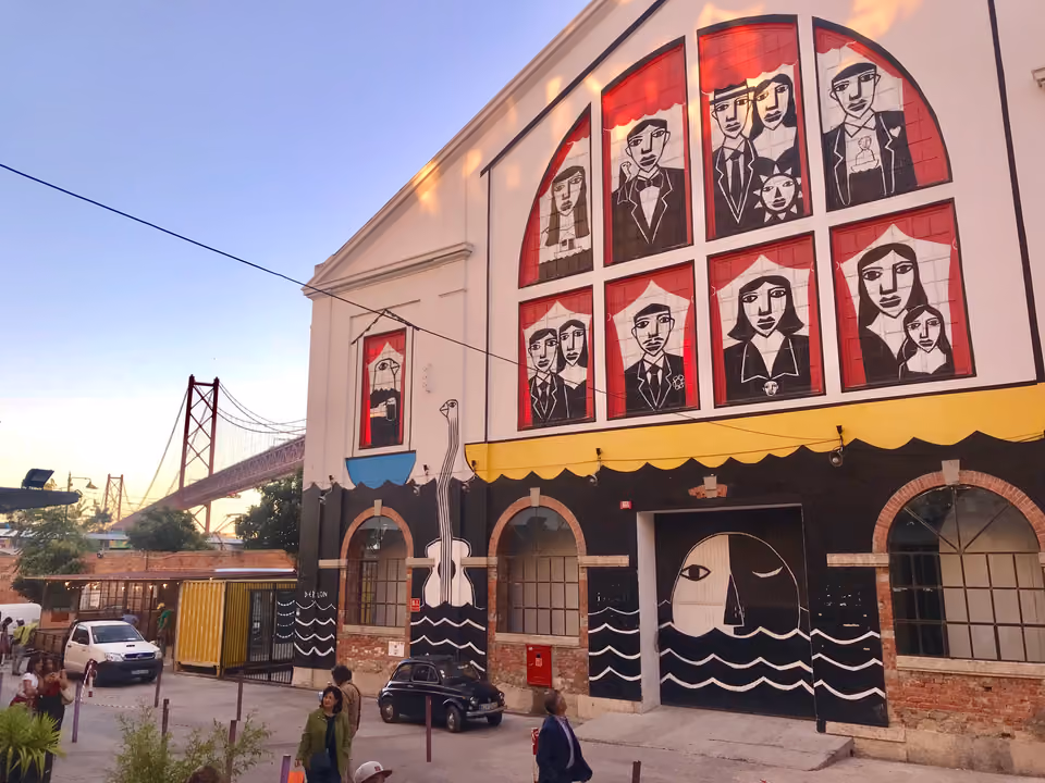 Fachada de um galpão da LX Factory em Lisboa, com grandes janelas em arco cobertas de murais figurativos, sob a Ponte 25 de Abril ao fundo