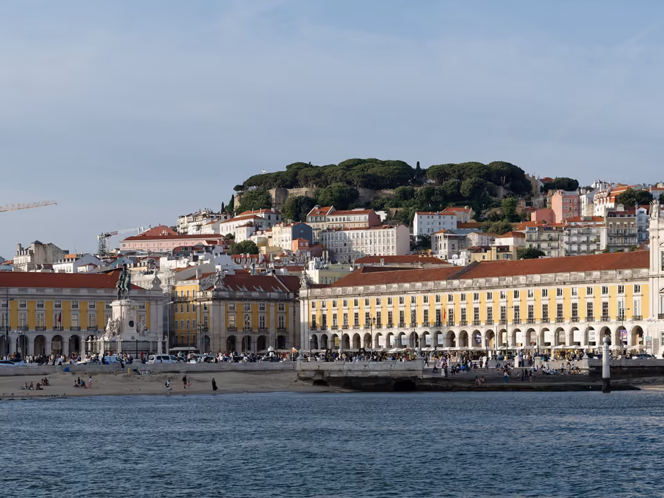 Praça do Comércio de Lisboa vista do Rio Tejo, com os edifícios amarelos em arcada, a estátua equestre de D. José e o Castelo de São Jorge na colina ao fundo
