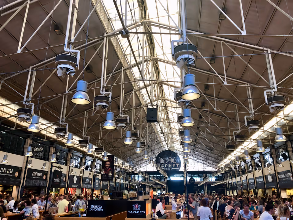 Salão do Time Out Market em Lisboa, sob a estrutura de ferro e vidro do antigo Mercado da Ribeira, com o letreiro central e as bancas cheias