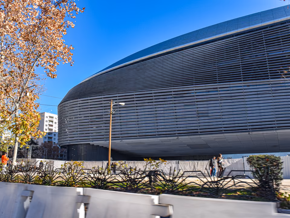 Fachada externa reformada do Estádio Santiago Bernabéu em Madri, o revestimento metálico curvo e ondulado erguido sobre a Castellana, com uma árvore de outono à esquerda