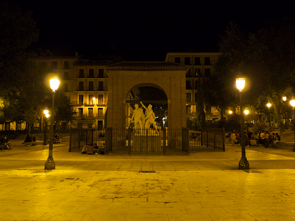 Plaza del Dos de Mayo, no bairro de Malasaña em Madri, à noite: o arco do antigo quartel de Monteleón com o monumento a Daoíz e Velarde, sob os lampiões, com gente sentada pela praça
