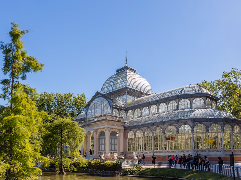 Palacio de Cristal do Parque del Retiro em Madri, pavilhão de vidro e ferro à beira do laguinho, cercado de árvores sob céu azul