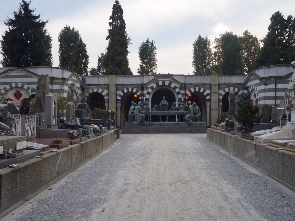 Alameda de saibro no Cimitero Monumentale de Milão levando ao túmulo da família Campari, um grupo escultórico em bronze diante da arcada listrada do cemitério
