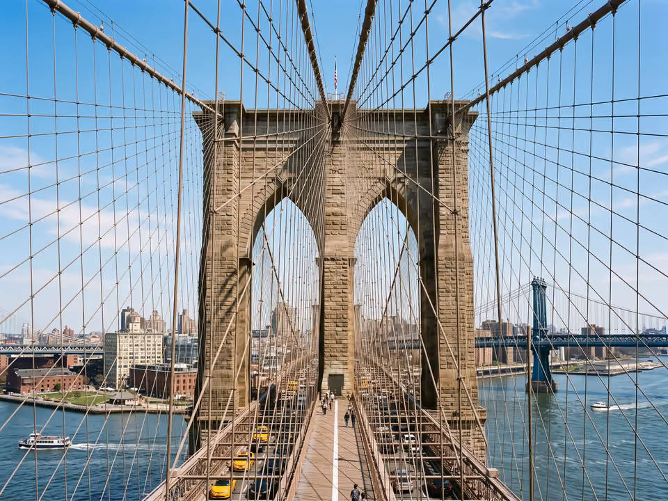 Torre de pedra da Ponte do Brooklyn vista da água, com os cabos de suspensão e o DUMBO ao fundo