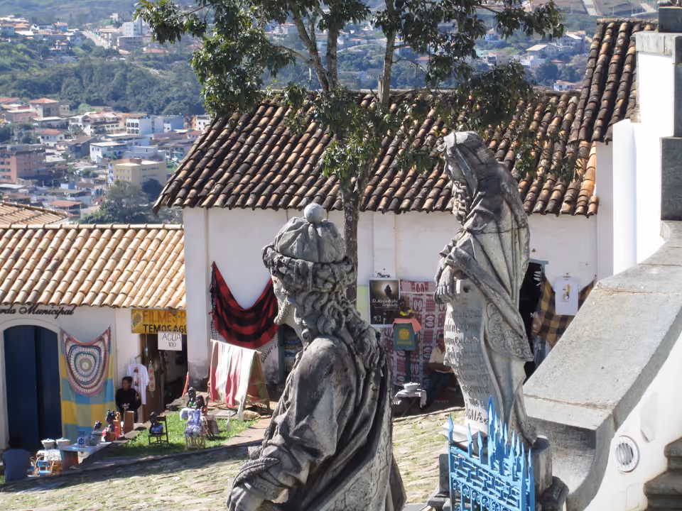 Duas das esculturas dos profetas de Aleijadinho no adro do Santuário do Bom Jesus de Matosinhos, em Congonhas, com a cidade e os telhados coloniais espalhados na encosta abaixo