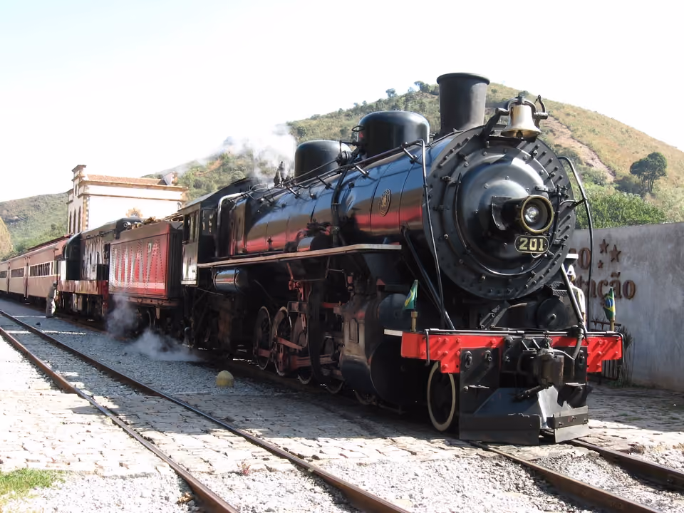 Locomotiva a vapor do Trem da Vale na estação, com fumaça saindo da chaminé e os vagões vermelhos vintage atrás, morros ao fundo