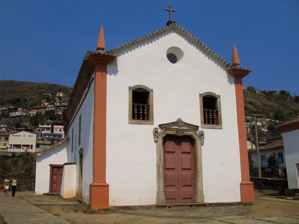 Capela do Padre Faria em Ouro Preto, pequena fachada branca com pilastras vermelhas, óculo circular e porta de madeira, sob céu azul