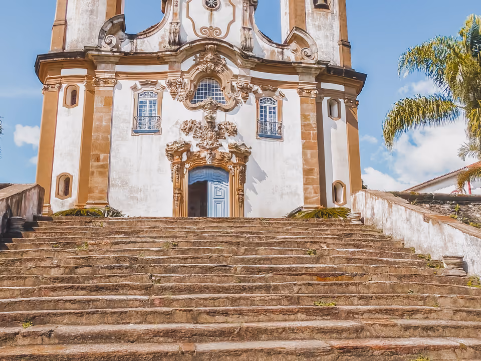 Portal rococó da Igreja de Nossa Senhora do Carmo em Ouro Preto, fachada branca com talha de pedra em torno da porta azul, no alto de uma larga escadaria de pedra