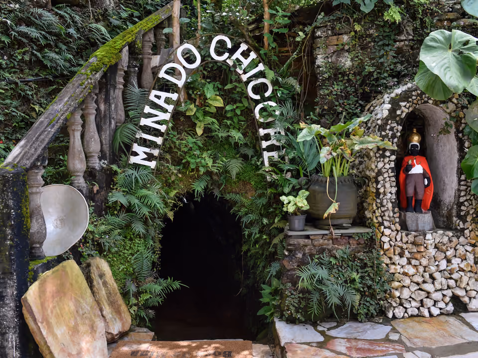 Entrada da Mina do Chico Rei em Ouro Preto, arco de pedra coberto de samambaias e musgo com o nome do local, e um nicho com a imagem de Chico Rei ao lado
