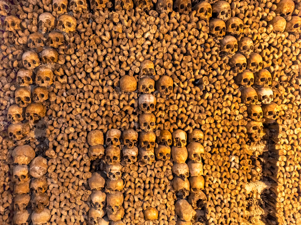 Parede das Catacumbas de Paris feita de crânios e fêmures empilhados em padrão, com uma cruz formada por caveiras ao centro