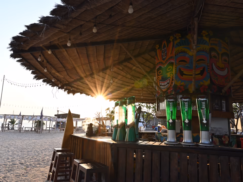 Bar de praia com cobertura de palha, torneiras de chope e máscaras coloridas, ao pôr do sol