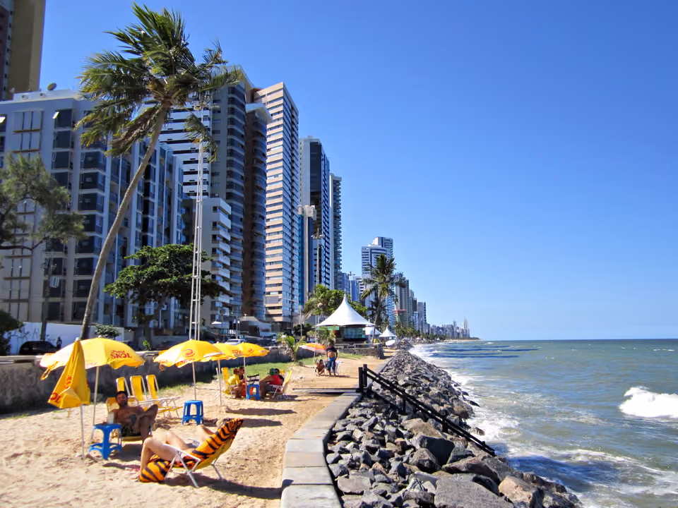 Orla da Praia de Boa Viagem em Recife, com coqueiros, guarda-sóis na areia, os prédios da avenida à beira-mar e a linha de recifes ao fundo