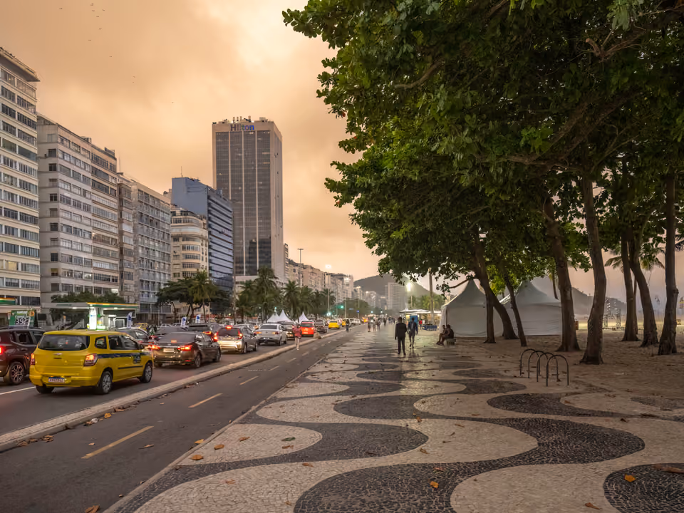 O calçadão de Copacabana com o mosaico de ondas em pedra portuguesa de Burle Marx ao entardecer, a Avenida Atlântica com trânsito à esquerda e a orla de prédios ao fundo