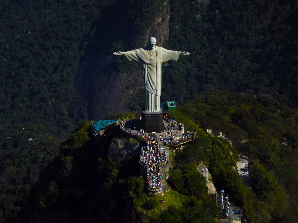 Estátua do Cristo Redentor de braços abertos no alto do Corcovado, sobre o pedestal e o mirante cheio de visitantes, cercada pela Floresta da Tijuca
