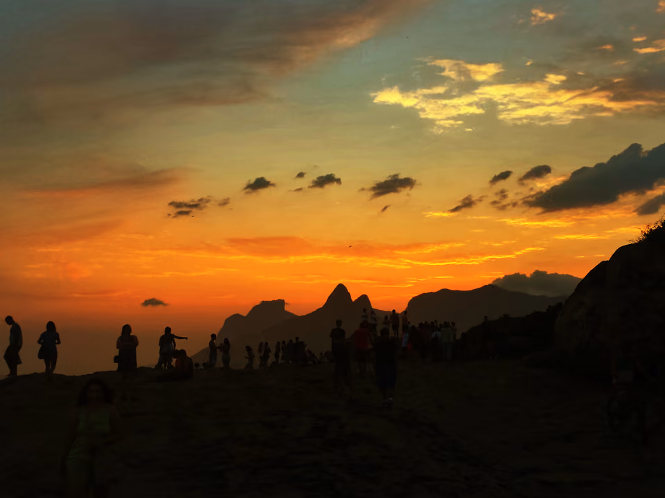 Pôr do sol visto da Pedra do Arpoador, com a silhueta do Morro Dois Irmãos e da Pedra da Gávea recortada contra o céu alaranjado e a multidão observando em contraluz