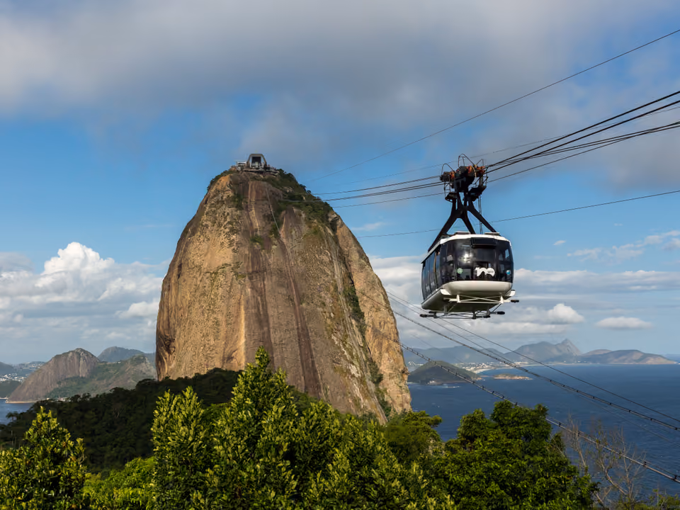 Morro do Pão de Açúcar com a estação no topo e um bondinho deslizando no cabo em direção ao cume, com a Baía de Guanabara e os morros de Niterói ao fundo