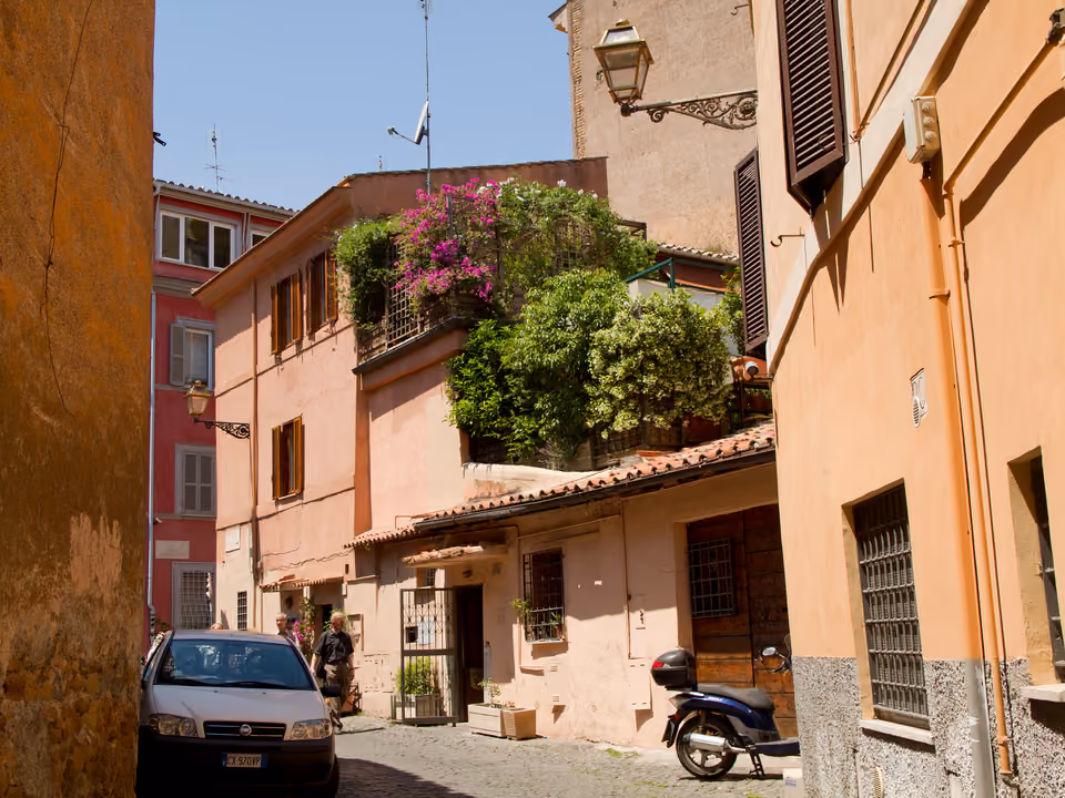 Vicolo estreito em Trastevere, Roma, com fachadas em tons terrosos e uma sacada coberta de trepadeiras floridas sobre a rua