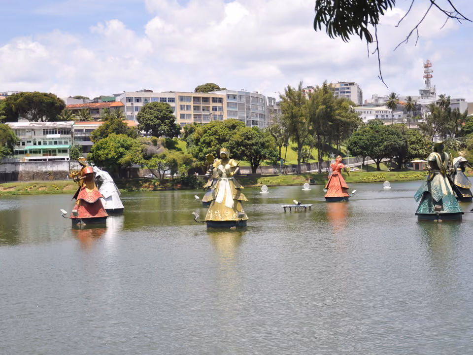 Esculturas dos orixás de Tatti Moreno erguidas sobre a água do Dique do Tororó em Salvador, figuras de braços abertos em túnicas coloridas, com a cidade ao fundo