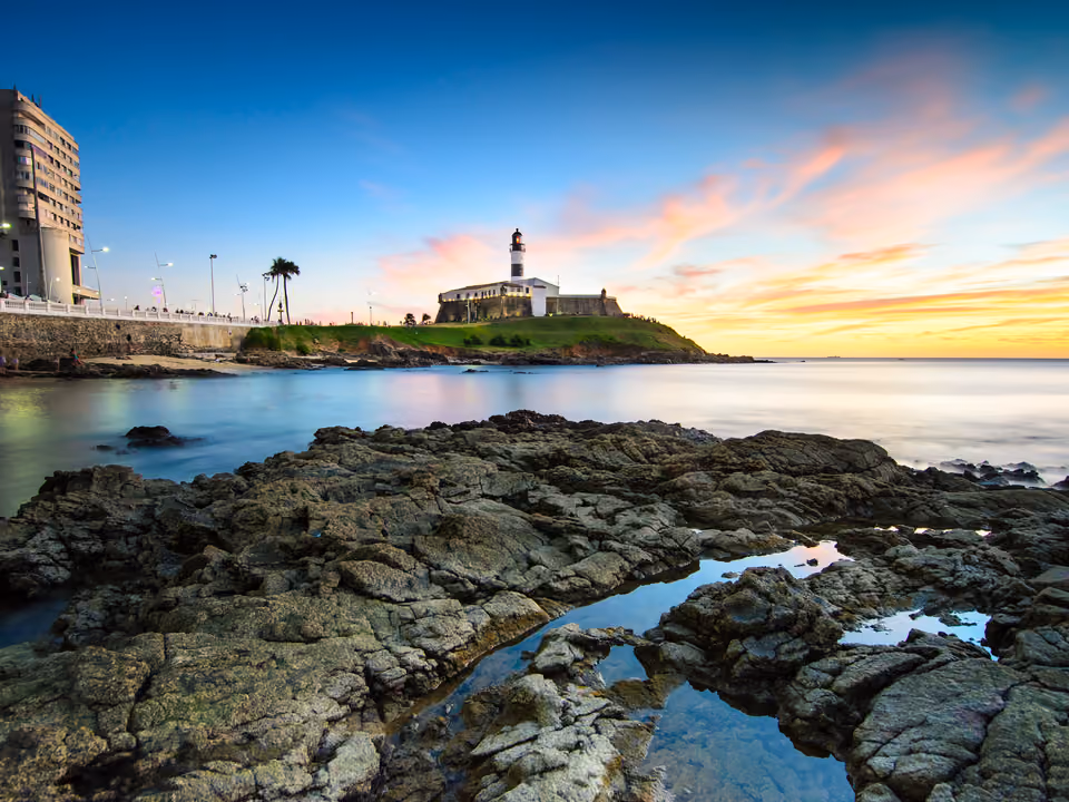 Farol da Barra em Salvador, a torre listrada de preto e branco sobre o Forte de Santo Antônio da Barra, à beira-mar ao pôr do sol, com poças entre as pedras em primeiro plano