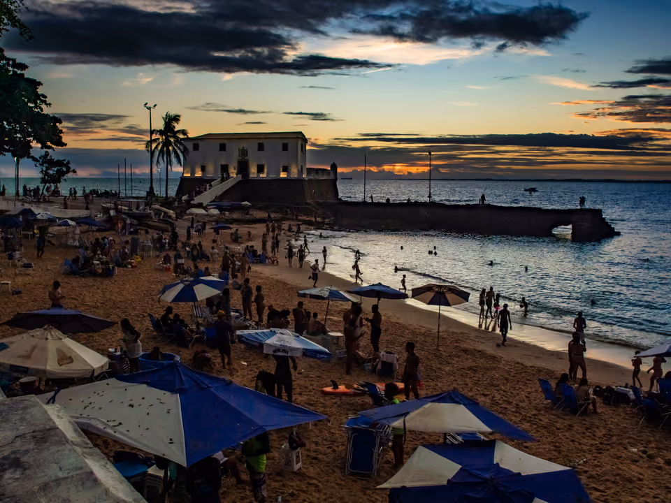 Praia do Porto da Barra em Salvador ao pôr do sol, a faixa curta de areia lotada de guarda-sóis e banhistas entre o Forte de Santa Maria e o mar