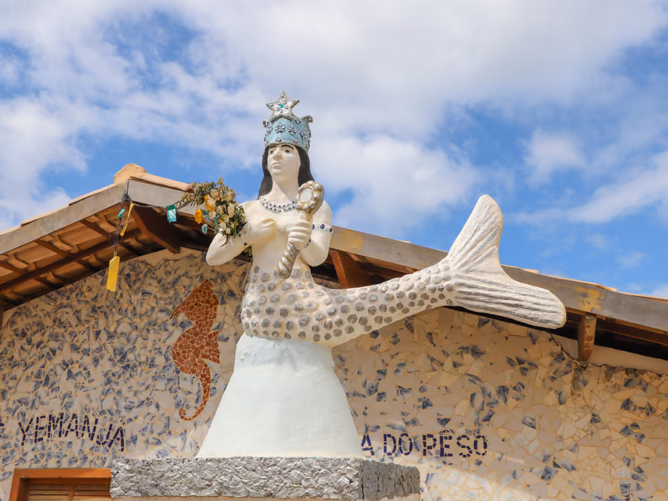 Escultura de Yemanjá — figura de sereia coroada segurando um espelho — no telhado da Casa de Iemanjá no Rio Vermelho, em Salvador, sobre a parede de mosaico com o nome 'Yemanjá'