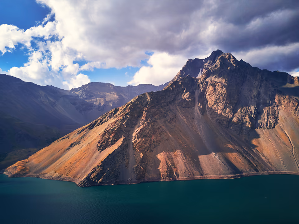 Águas turquesa do Embalse El Yeso no Cajón del Maipo, ao pé de um pico árido dos Andes sob nuvens dramáticas