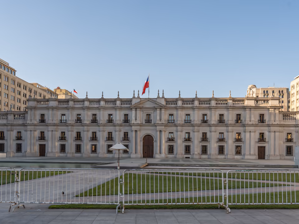 Fachada neoclássica do Palácio de La Moneda em Santiago, com colunas e a bandeira do Chile hasteada, vista da praça