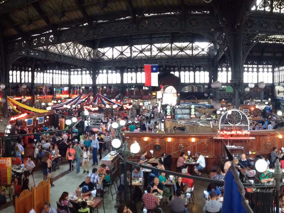 Interior do Mercado Central de Santiago visto do alto, com a estrutura de ferro do teto, a bandeira do Chile e os restaurantes de frutos do mar cheios no almoço