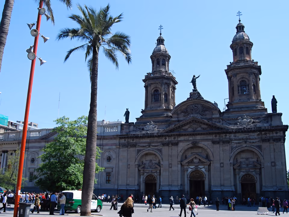 Catedral Metropolitana de Santiago vista da Plaza de Armas, com as duas torres, uma palmeira alta e a praça cheia de gente