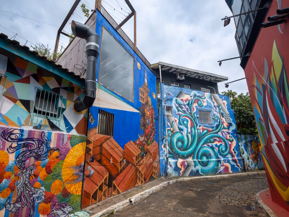 A ruela de paralelepípedo do Beco do Batman, na Vila Madalena, em São Paulo, curvando entre prédios com as paredes inteiras cobertas de grafites coloridos