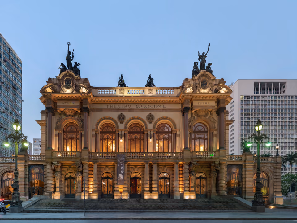Fachada do Theatro Municipal de São Paulo ao anoitecer, o prédio eclético de 1911 iluminado de dourado, com as esculturas no telhado e os lampiões de ferro na frente